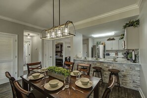 A dining area with a wooden table set for four, a hanging light fixture, and a kitchen in the background. -         MOUNTAIN VIEW BLESSINGS VIEW SERENITY of MTS. Newly Remodeled 2Bed2Bath (Pigeon Forge)