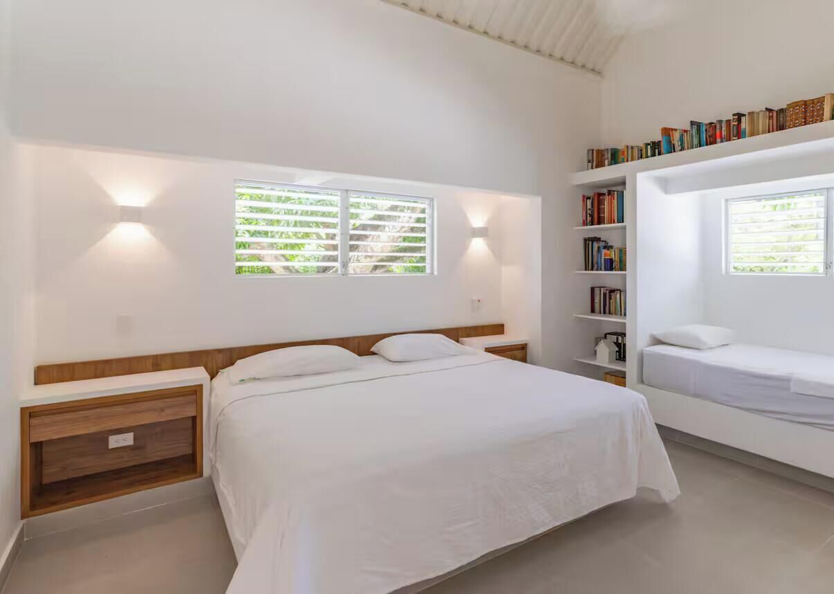A modern bedroom with a large bed, bedside tables, a bookshelf, and a window with blinds.