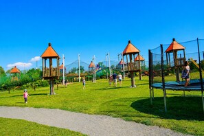 Ein Spielplatz mit verschiedenen Spielgeräten, darunter Schaukeln und Kletterbereichen, umgeben von einer grünen Wiese.