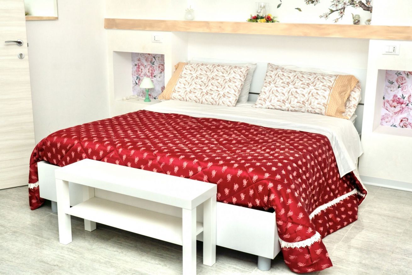 Ein Bett mit einer rot gemusterten Tagesdecke, weißen Kissen und einer weißen Bank davor.