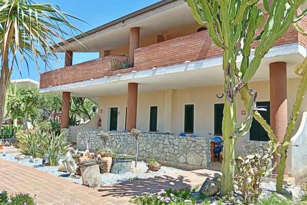 A two-story building with a balcony, stone walls, and a variety of plants. - This charming and tastefully furnished apartment, approximately 60 m², is located on the ground or first floor of ** Residence**, just 600 metres from the beach.The apartment features a welcoming living room with a sofa, a fully equipped kitchen, a bath (Ricadi)
