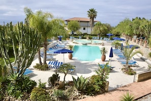 A pool area with lounge chairs and umbrellas, surrounded by cacti and palm trees. - This charming and tastefully furnished apartment, approximately 60 m², is located on the ground or first floor of ** Residence**, just 600 metres from the beach.The apartment features a welcoming living room with a sofa, a fully equipped kitchen, a bath (Ricadi)