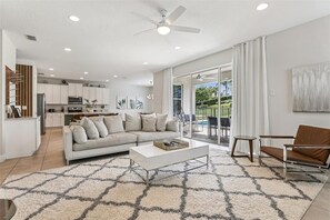 A modern living room with a sofa, coffee table, and a view of a patio with outdoor furniture. - Orlando Home: Private Pool & Themed Rooms (Kissimmee)
