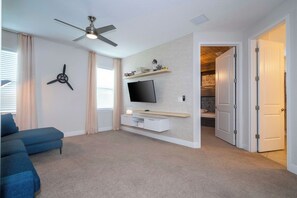 A living room with a ceiling fan, a flat-screen TV mounted on the wall, a blue sofa, and a small table with decorative items. - Pool, Spa & Game Room Home for 12 at Encore (Kissimmee)