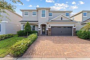 A two-story house with a brick driveway, a garage, and a well-maintained front yard with shrubs and a small tree. - Orlando Home: Private Pool & Themed Rooms (Kissimmee)