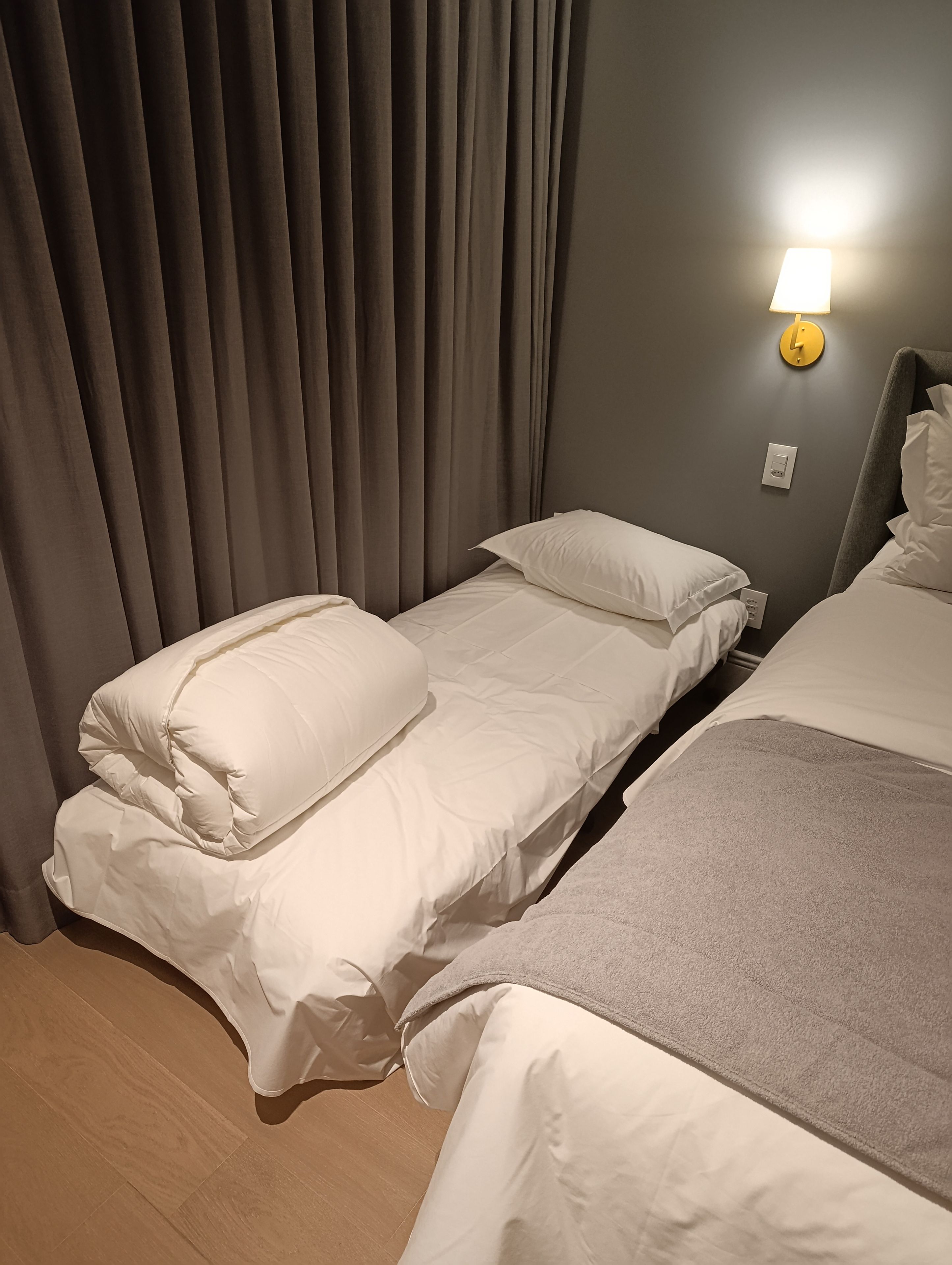 Habitación de hotel con dos camas individuales, una cortina gris, una lámpara fijada a la pared y un termostato.
