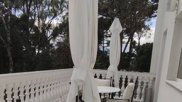 An outdoor patio area with white tables and chairs, umbrellas, and a view of trees and a building in the background.