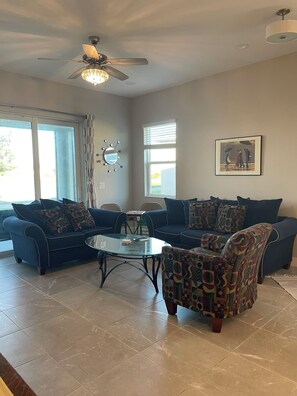 A living room with a ceiling fan, a glass coffee table, two sofas, and patterned cushions. - Golf Course-Pond, 2 Bed/2 Bath+ Den 2022 Villa-Quiet Active 55+ Gated Community (Port Charlotte)