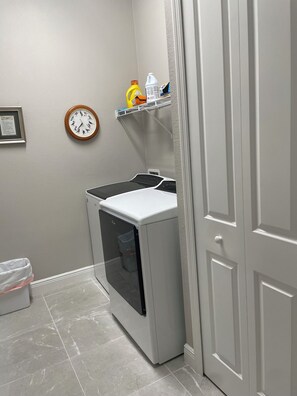 A laundry room with a front-loading washer and dryer, a clock, and a shelf with cleaning supplies. - Golf Course-Pond, 2 Bed/2 Bath+ Den 2022 Villa-Quiet Active 55+ Gated Community (Port Charlotte)