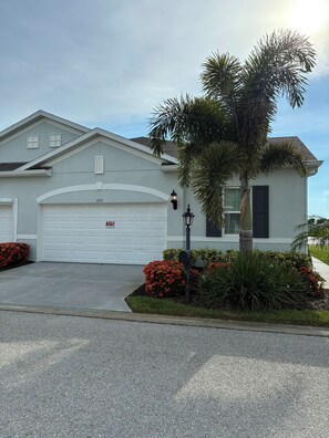 A two-story residential building with a white garage door, a small porch with a lamp, and a palm tree. - Golf Course-Pond, 2 Bed/2 Bath+ Den 2022 Villa-Quiet Active 55+ Gated Community (Port Charlotte)