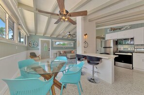 Eine geräumige Küche mit modernen Geräten, ein Essbereich mit einem Glastisch und türkisfarbenen Stühlen sowie ein Deckenventilator.