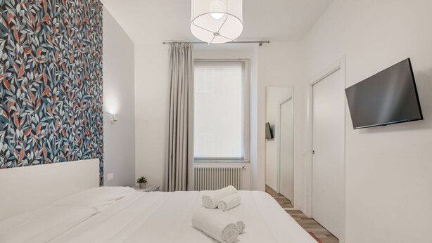 Hotellihuone, jossa on sänky, televisio, ikkuna verhoilla ja kuvioitu seinä.