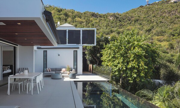 A modern house with a glass infinity pool, outdoor seating, and a lush green hillside in the background.