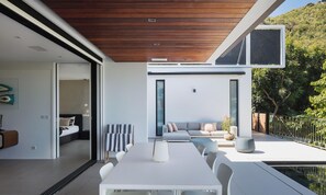 A modern outdoor dining area with a pool, a seating area with a sofa and cushions, and a view of a lush garden.