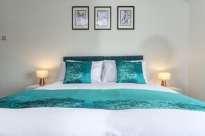 A bed with a teal and white bedspread, two white pillows, and a teal headboard. There are two bedside lamps on either side of the bed. Above the bed, there are three framed artworks on the wall. - The Waltons – Ideal Durham Escape - Nature Lovers - Pass the Keys (New Brancepeth)