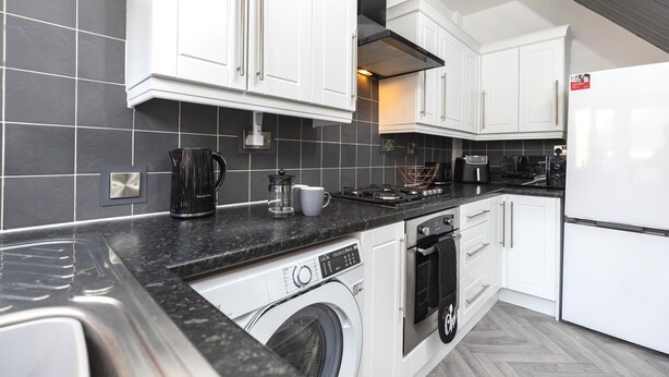 A modern kitchen with white cabinets, a black countertop, a built-in washing machine, and a stainless steel range hood. - The Waltons – Ideal Durham Escape - Nature Lovers - Pass the Keys (New Brancepeth)