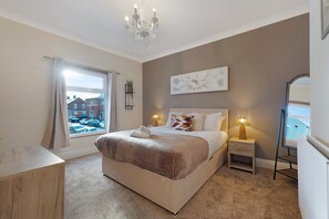 A bedroom with a large bed, bedside lamps, a chandelier, a window with curtains, and a view of buildings outside. - Characterful 3 Bed | Sleeps 6 | Central Darlington - Pass the Keys (Darlington)