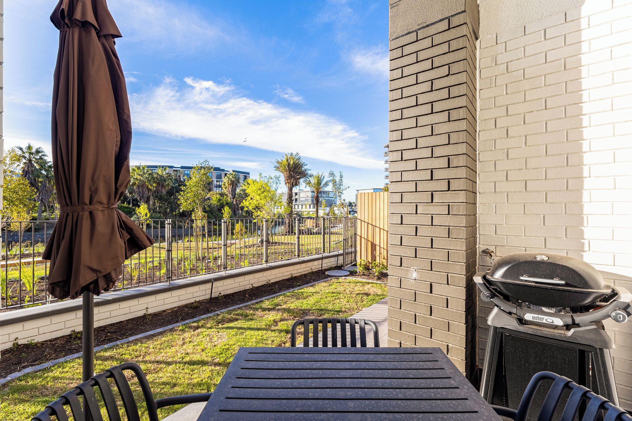 An outdoor patio with a table and chairs, a barbecue grill, and a view of a fenced garden area.