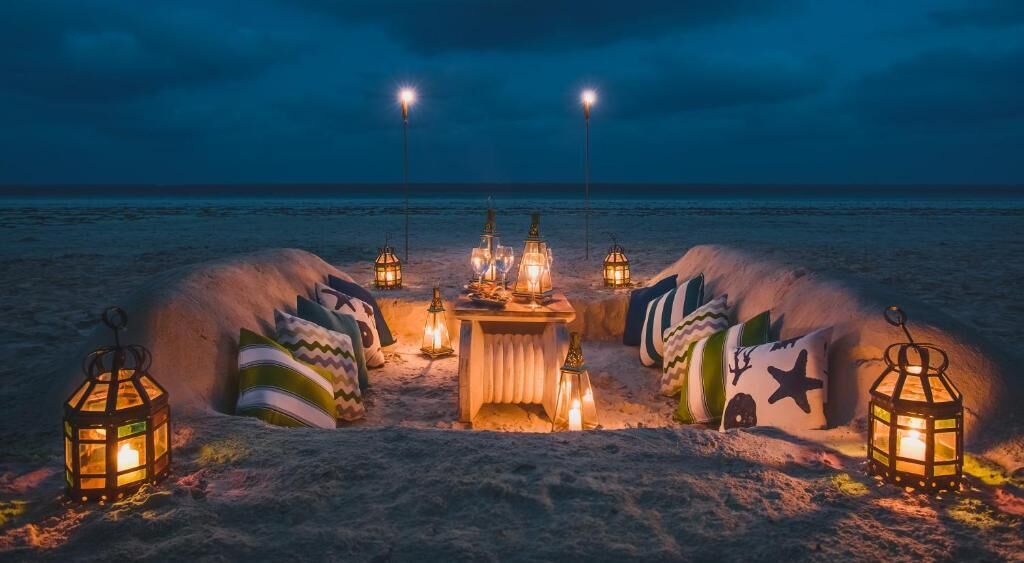 Un ambiente playero con una mesa dispuesta para dos personas, rodeada de faroles y cojines.