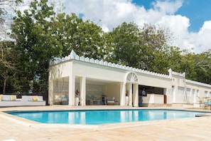 A poolside area with a covered seating area, a pool, and a building with a white facade and a decorative roof. - The Zubeida (Diani Beach)