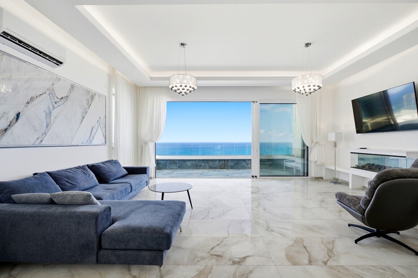 A modern living room with a sofa, a chair, a TV, and a view of the ocean.