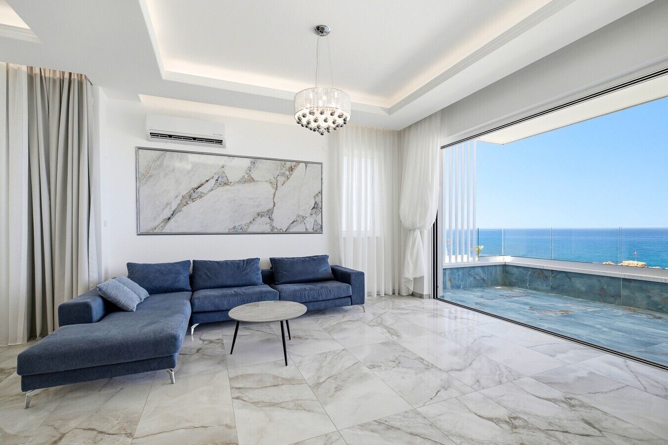 A modern living room with a sectional sofa, a round coffee table, and a large window offering a view of the sea.