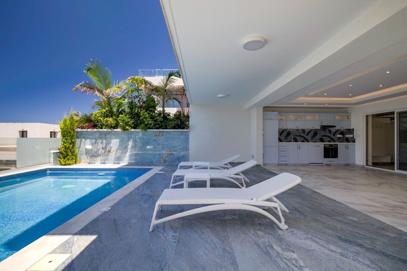 Un área de piscina exterior moderna con sillones de descanso, una cocina con salpicadero de mármol y un cielo azul despejado.