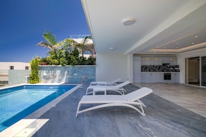 A modern outdoor pool area with lounge chairs, a kitchen with marble backsplash, and a clear blue sky. - Olymp Royal Palace - Ultra Luxury Beachfront House (Paphos)