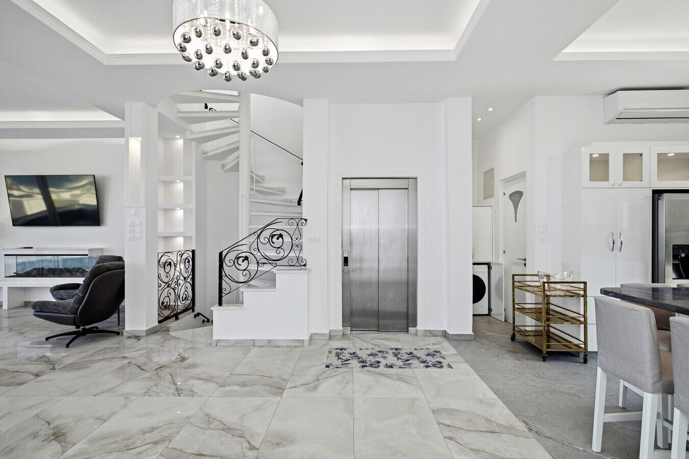A modern interior with a marble floor, a staircase with decorative railings, a dining area with a glass table, and a kitchen with white cabinets.