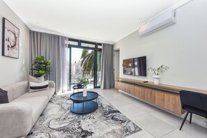 A modern living room with a sofa, a coffee table, a TV, and a view through a glass door. - The Paramount | Contemporary Luxury (Johannesburg)