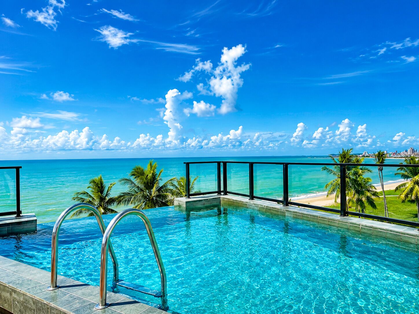 A clear blue sky, a swimming pool with a glass barrier, and a view of the ocean and beach.