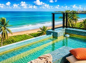 A pool with a view of a beach and ocean, surrounded by palm trees and a city skyline. - Breeze Residence 317 Beachfront Flat at Bessa (João Pessoa)