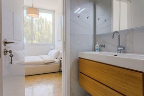 A modern bathroom with a large mirror, a sink, and a toilet. There is a window providing natural light and a view of trees outside. - Olymp Luxury Residence - Luxury Beachfront Apartment (Kissonerga)