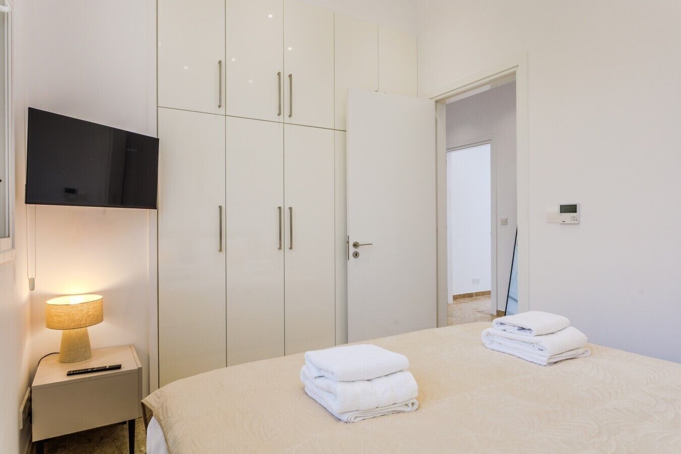 A bedroom with a bed, a TV mounted on the wall, a bedside lamp, and a door leading to another room.
