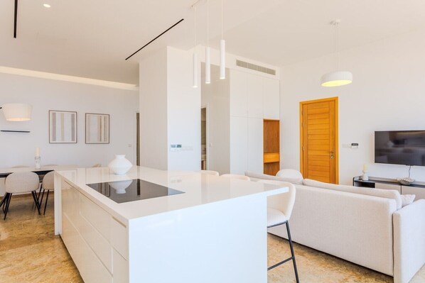 A modern living space with a central island, a sofa, a TV, and a dining area. - Olymp Luxury Residence - Luxury Beachfront Apartment (Kissonerga)