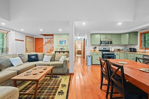 Un interior moderno con una sala de estar que incluye un sofá, una mesa de centro y un tapete estampado. Una zona de comedor con una mesa y sillas de madera está al lado de una cocina con armarios verdes y electrodomésticos modernos.
