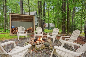 Un espace boisé avec un foyer entouré de fauteuils Adirondack, un abri à bois de chauffage et un drapeau.
