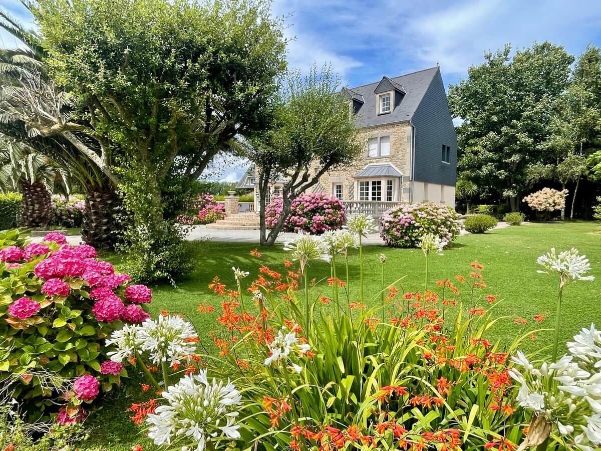 A house with a stone facade, surrounded by a well-maintained garden with various flowering plants and a lawn.