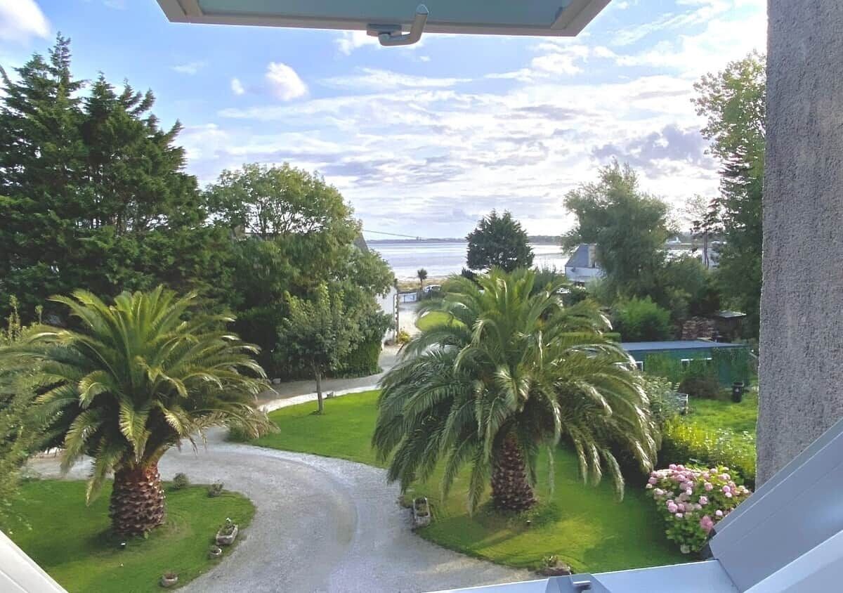 Une vue depuis un balcon donnant sur un jardin bien entretenu, avec des palmiers et un sentier menant à un plan d’eau.