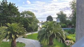 Une vue depuis un balcon donnant sur un jardin bien entretenu, avec des palmiers et un sentier menant à un plan d’eau.
