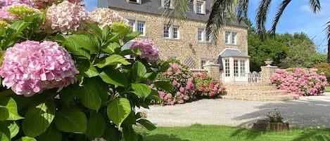 A stone building with a symmetrical facade, a central entrance, and a well-maintained garden with pink hydrangeas.