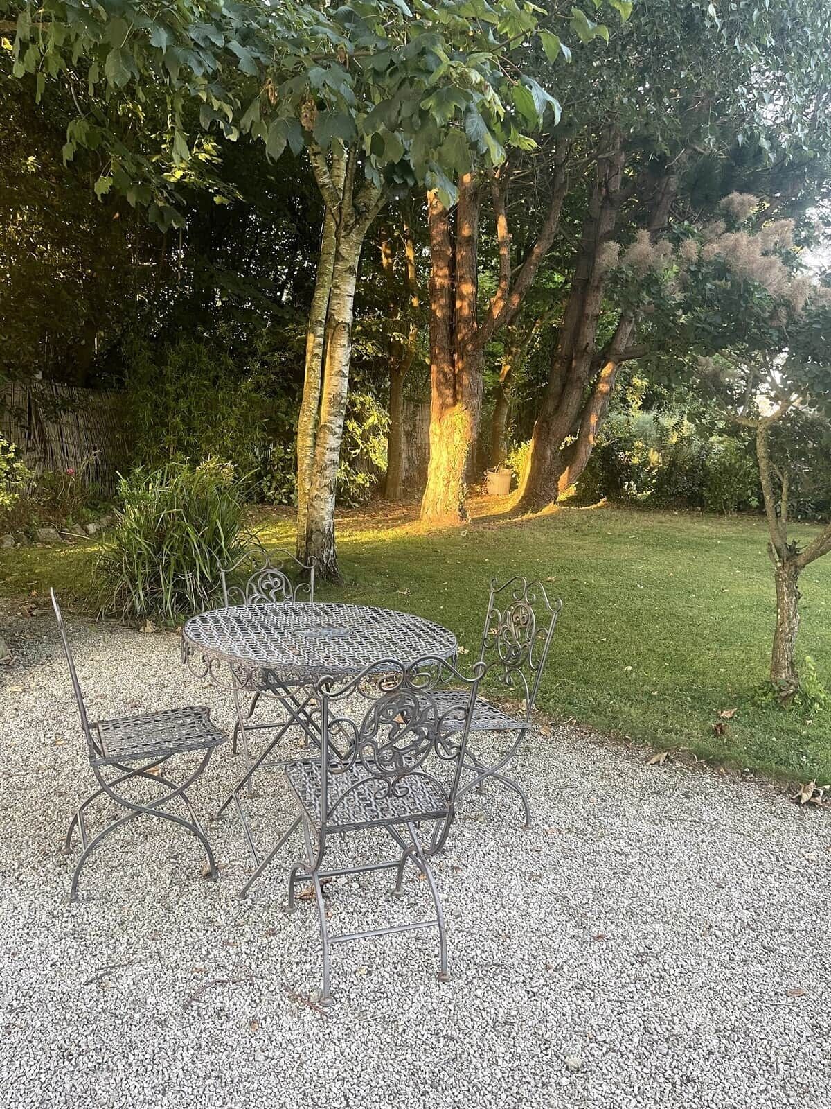 Een metalen tuinset met tafel en stoelen, geplaatst op een grindpad, omgeven door bomen en gras.