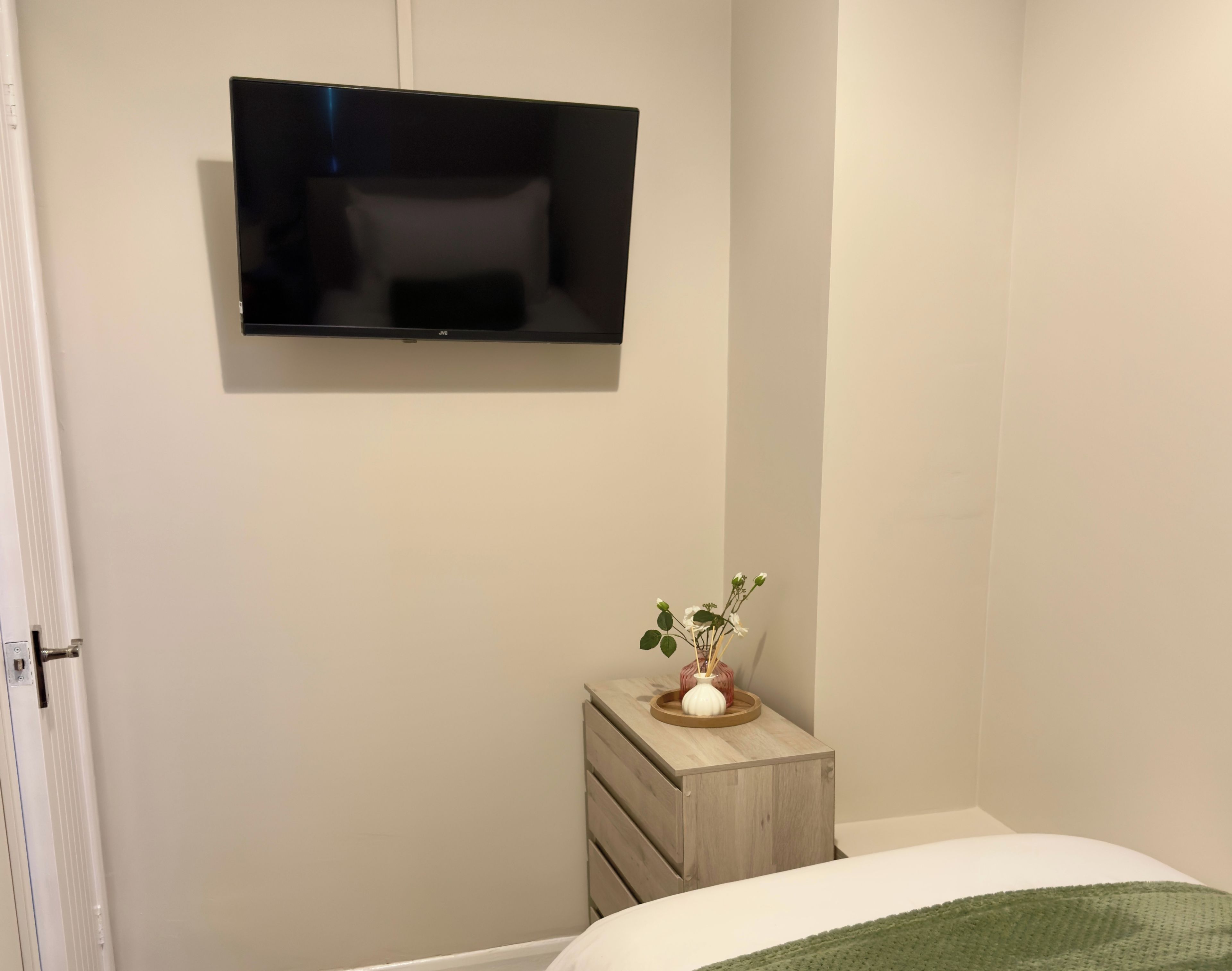 Ein Schlafzimmer mit einem Bett, einem Nachttisch mit Blumenvase, einer Kommode und einem an der Wand montierten Fernseher.