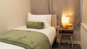 Una cama bien hecha con un almohadón verde y uno blanco, una mesita de noche con una lámpara, y un radiador en una habitación con paredes de tonos claros.