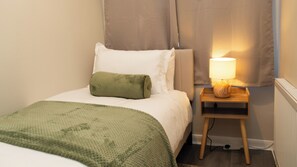 Una cama bien hecha con un almohadón verde y uno blanco, una mesita de noche con una lámpara, y un radiador en una habitación con paredes de tonos claros.