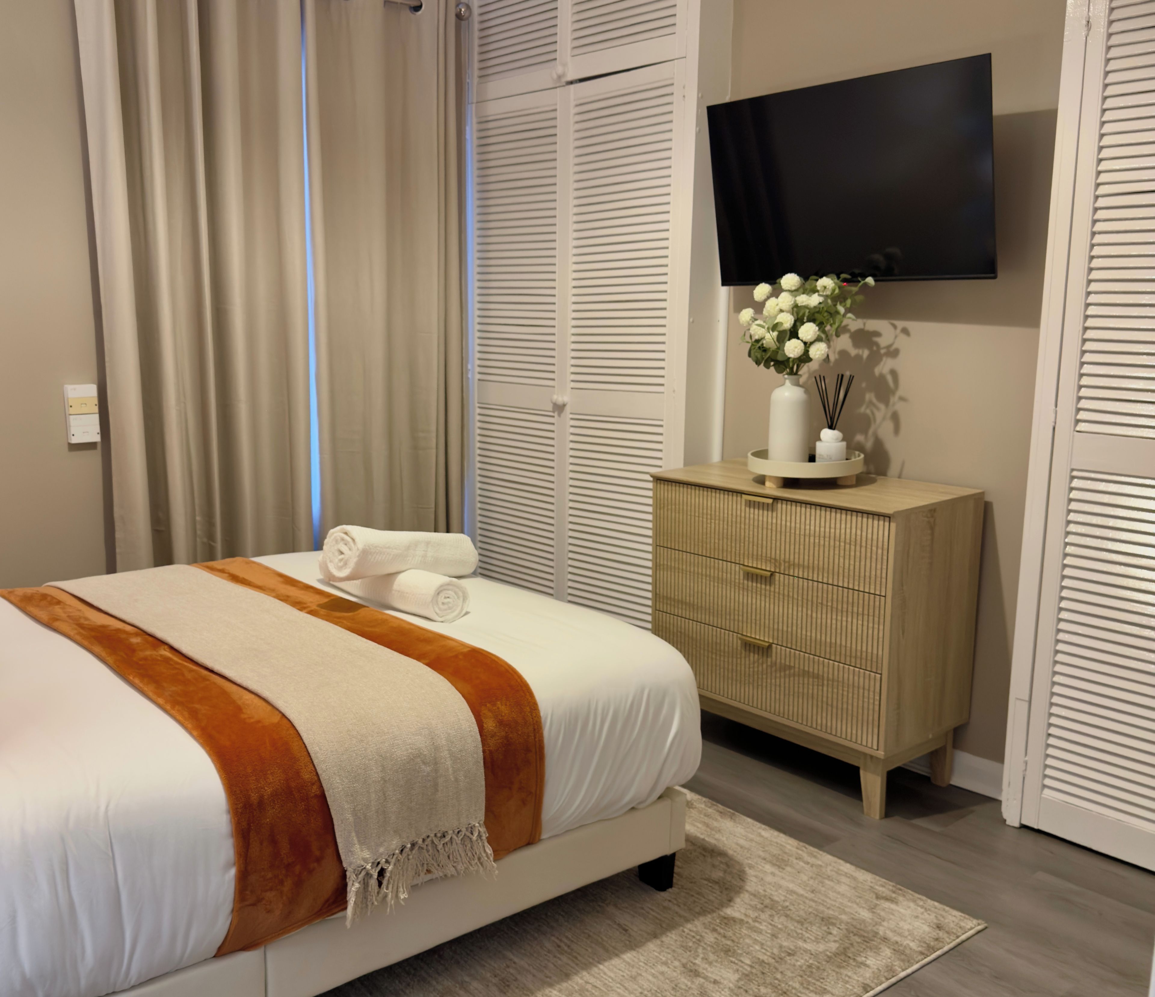 Cama bem arrumada com manta branca e marrom, cômoda de madeira com um vaso de flores e TV de tela plana na parede.