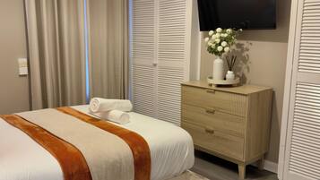 Cama bem arrumada com manta branca e marrom, cômoda de madeira com um vaso de flores e TV de tela plana na parede.