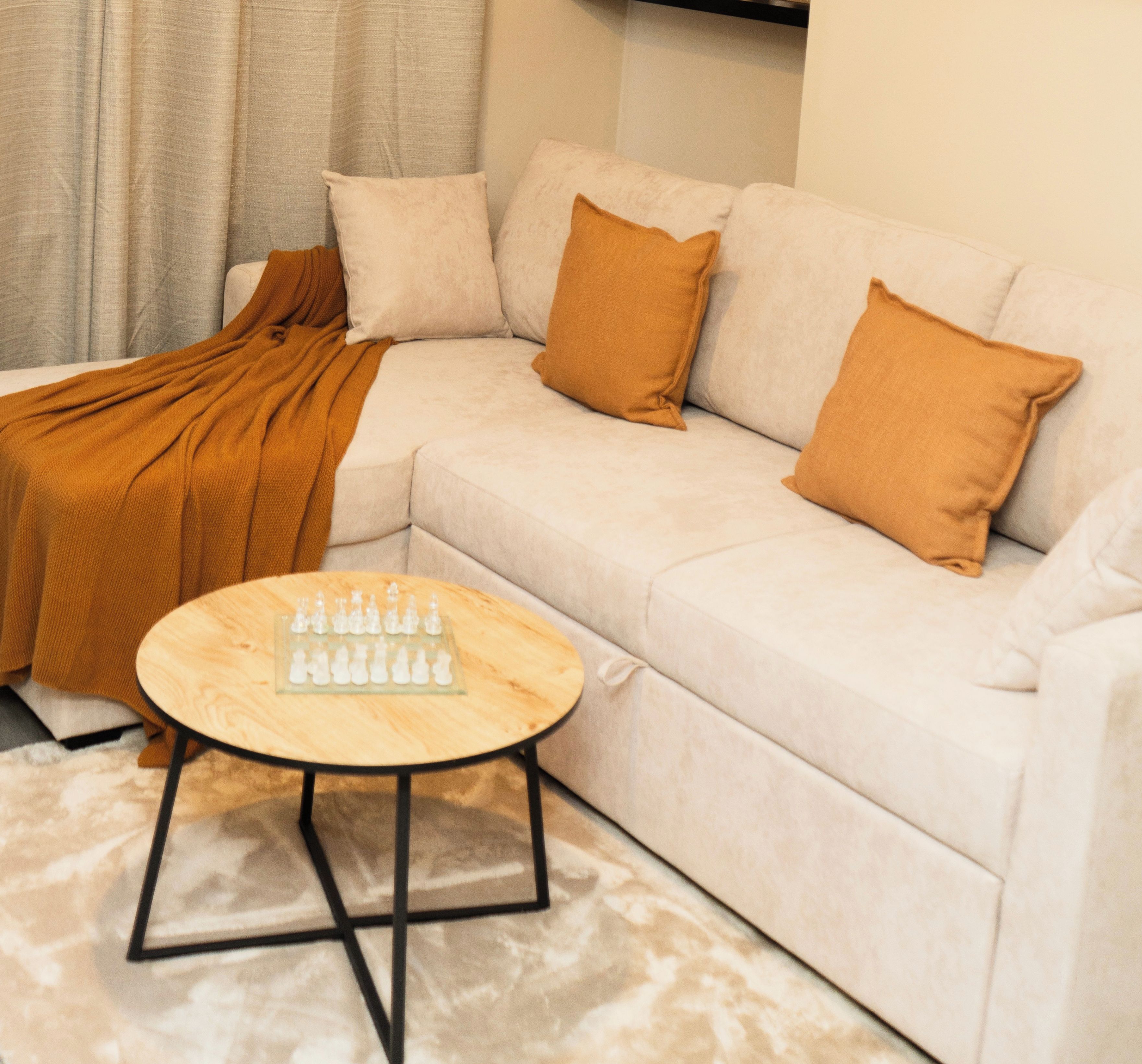 Uma sala de estar moderna com um sofá branco, duas almofadas marrons, uma mesa de centro de madeira com peças de xadrez e um tapete bege.