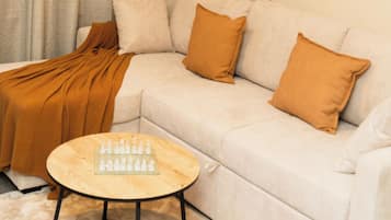 Uma sala de estar moderna com um sofá branco, duas almofadas marrons, uma mesa de centro de madeira com peças de xadrez e um tapete bege.