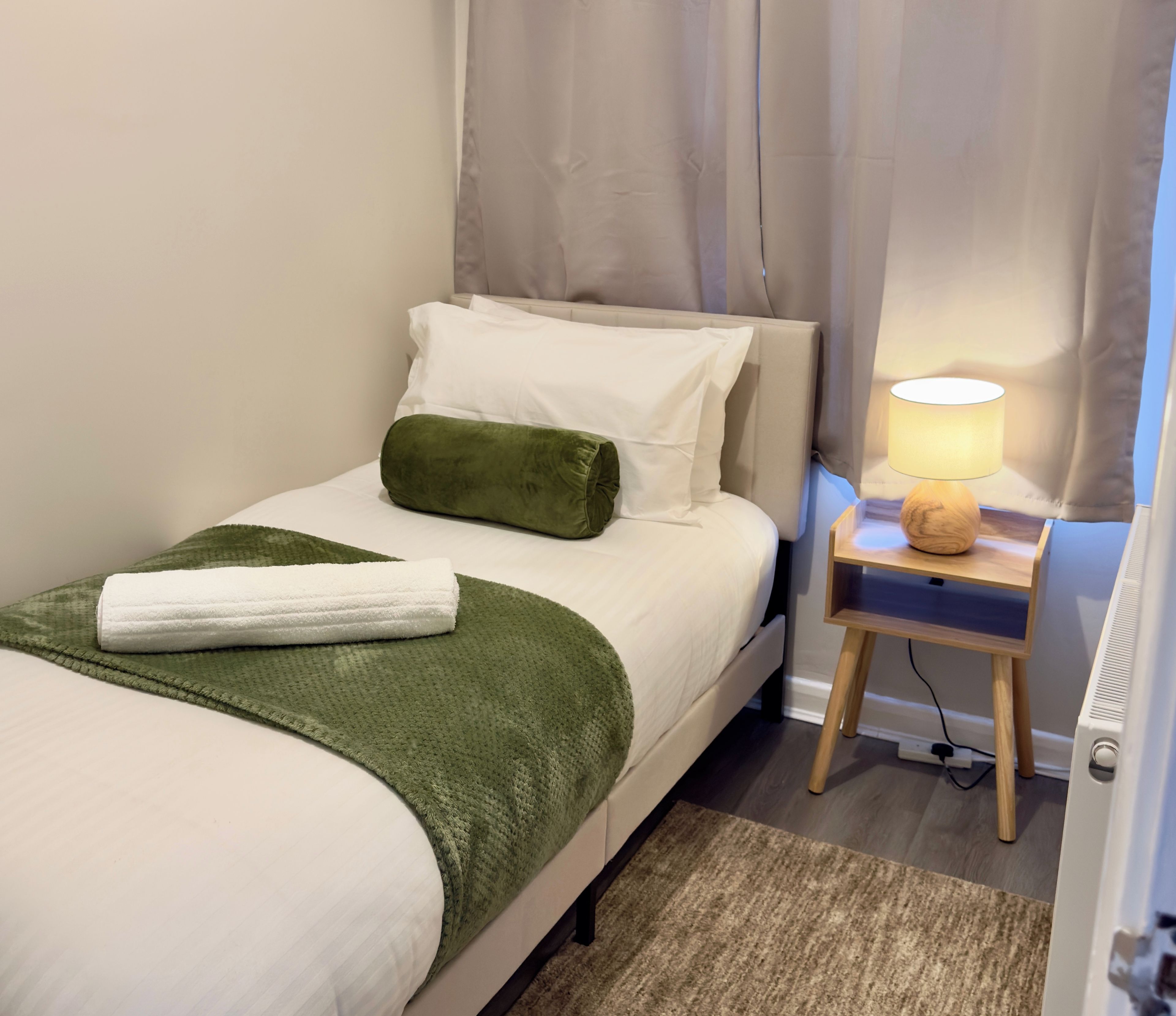 A neatly made bed with a green throw, a white towel, and a green pillow. There is a bedside table with a lamp and a small wooden stool.